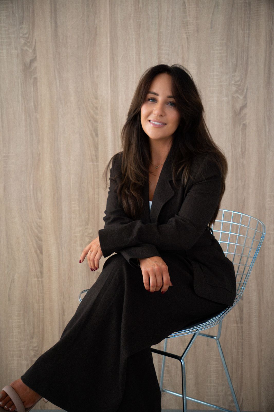Melissa, founder of Savoir, sitting on a wire chair in a dark brown suit, smiling