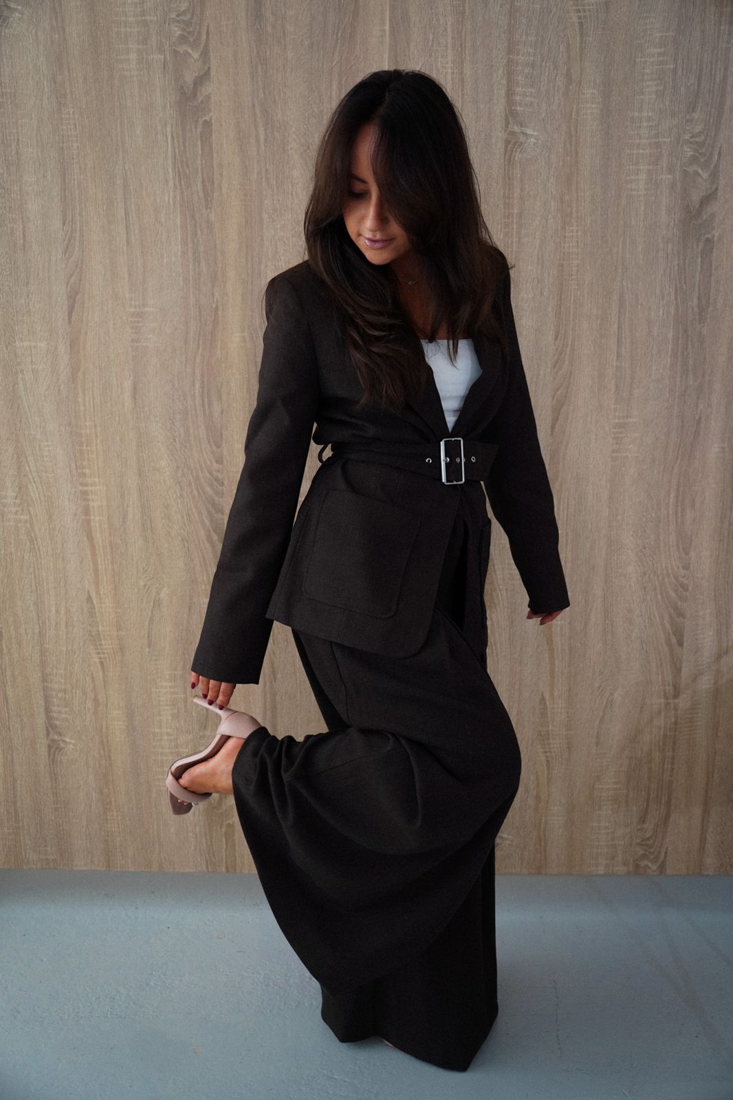 Melissa standing in a belted brown suit, holding one heel and looking down with a smile